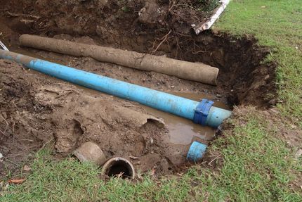 Exposed blue and brown pipes in a muddy trench beside grass and a small drainage opening