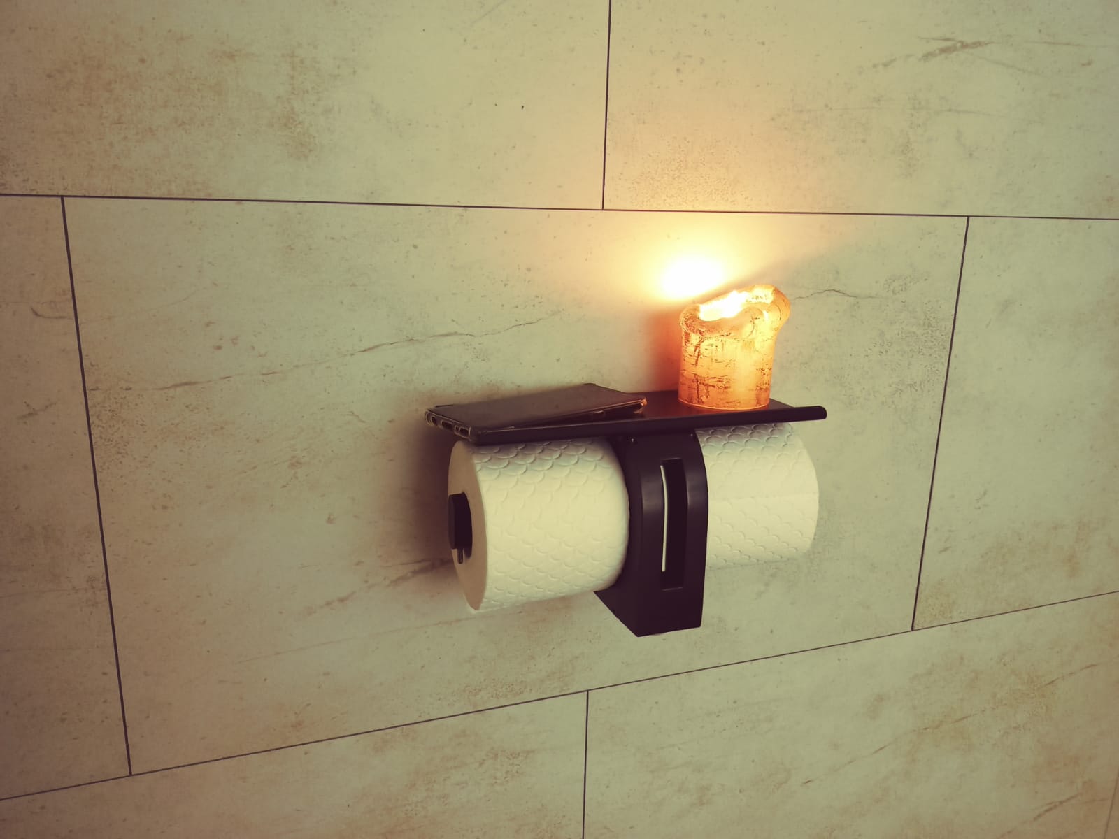 Wall-mounted toilet paper holder with a lit candle on top, against a tiled wall.