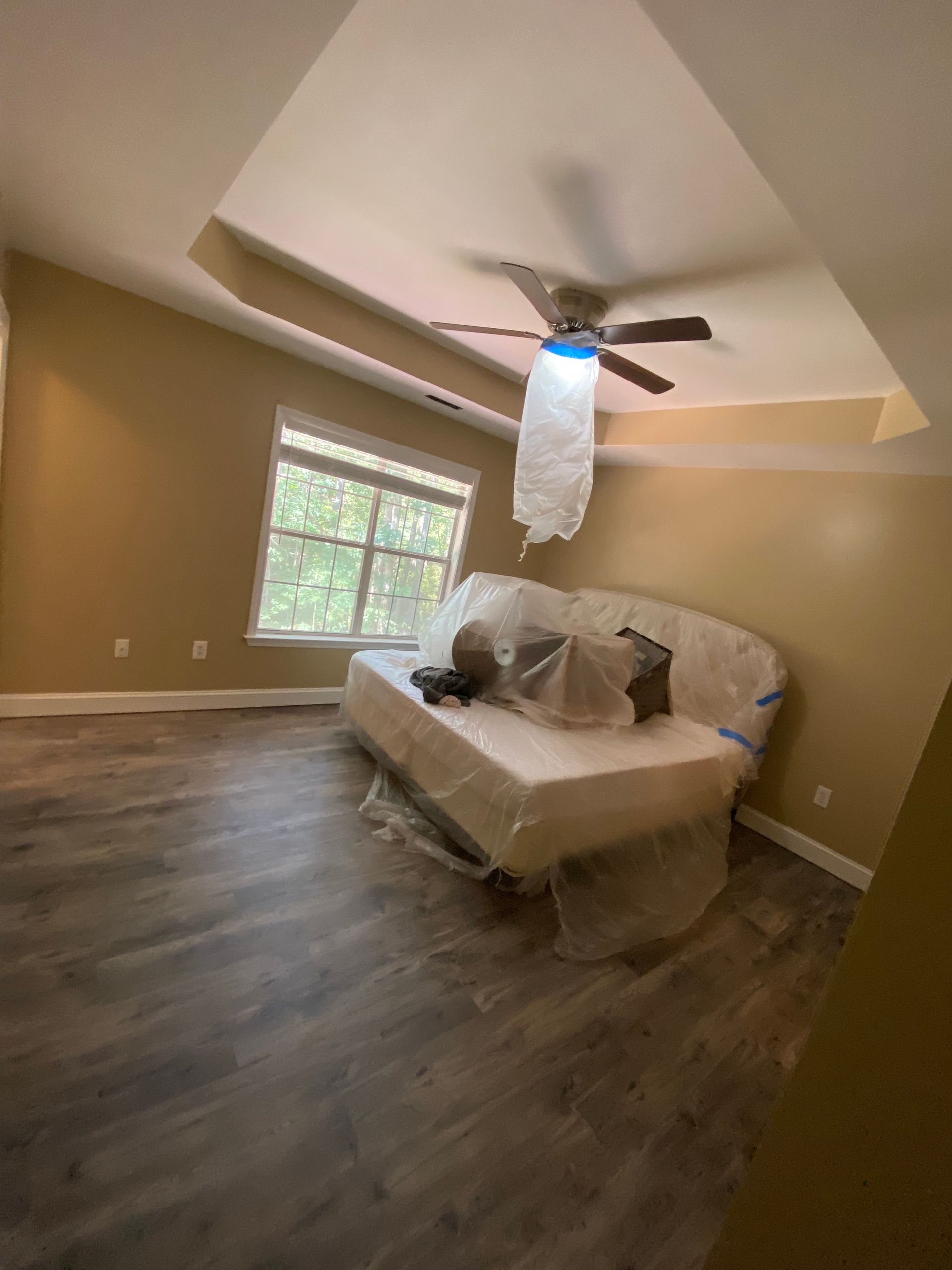 Bedroom with tan walls, dark wood floor, and a bed covered in plastic near a window. Ceiling fan visible.