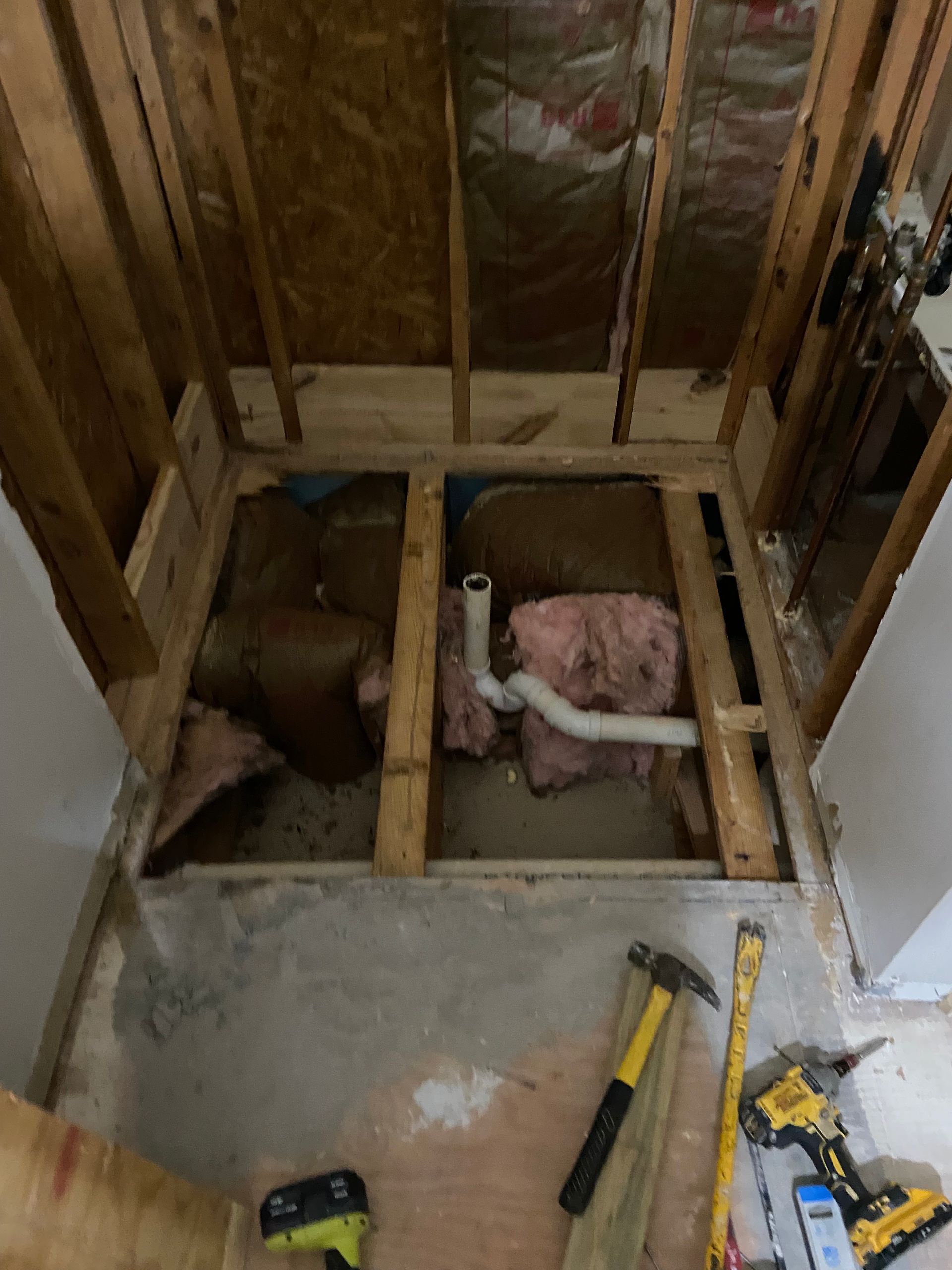 Bathroom under construction. Exposed framing, insulation, plumbing, and tools on floor.