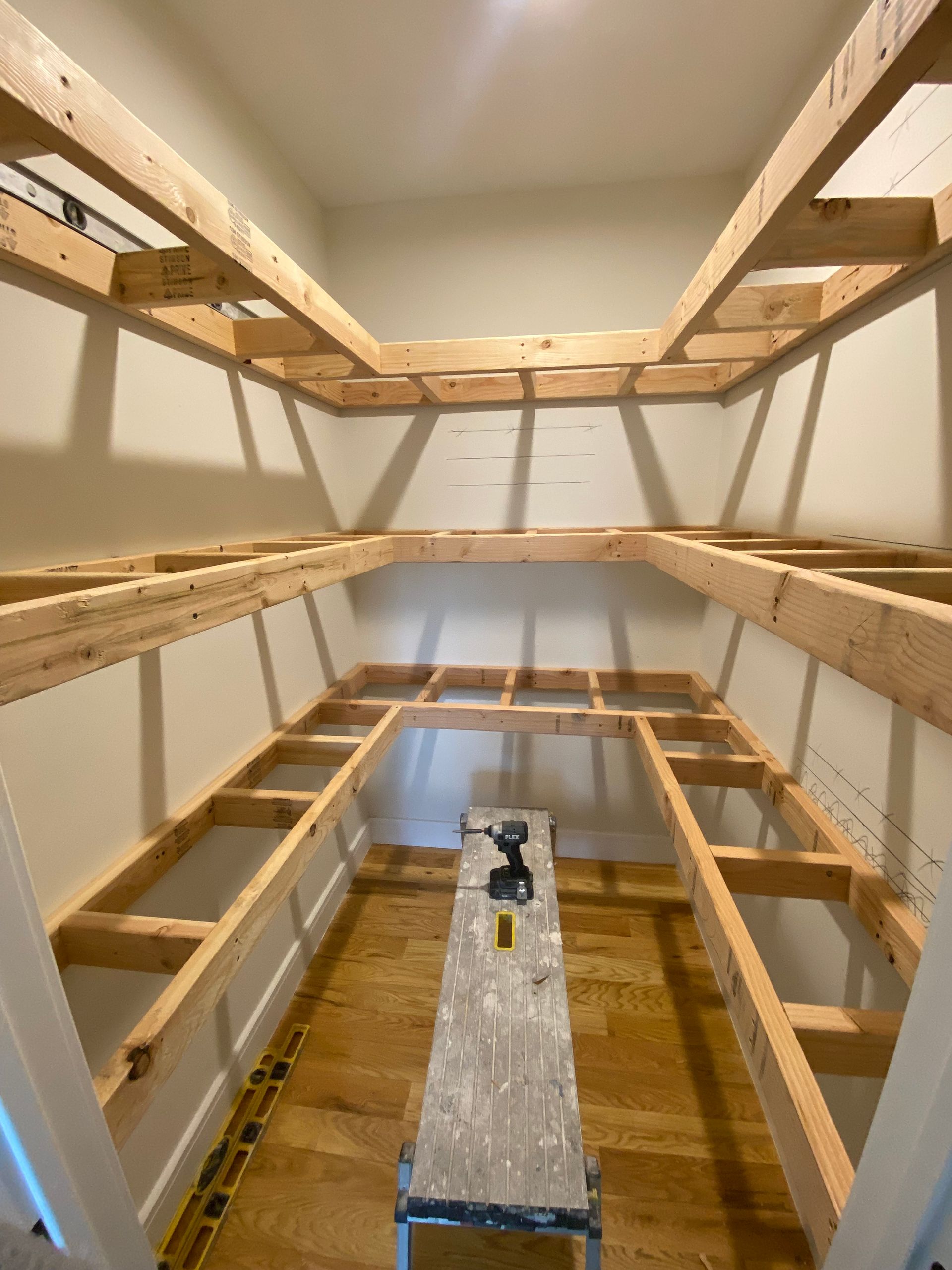 Wooden shelving framework installed inside a pantry. A stepladder with a drill sits in the center.