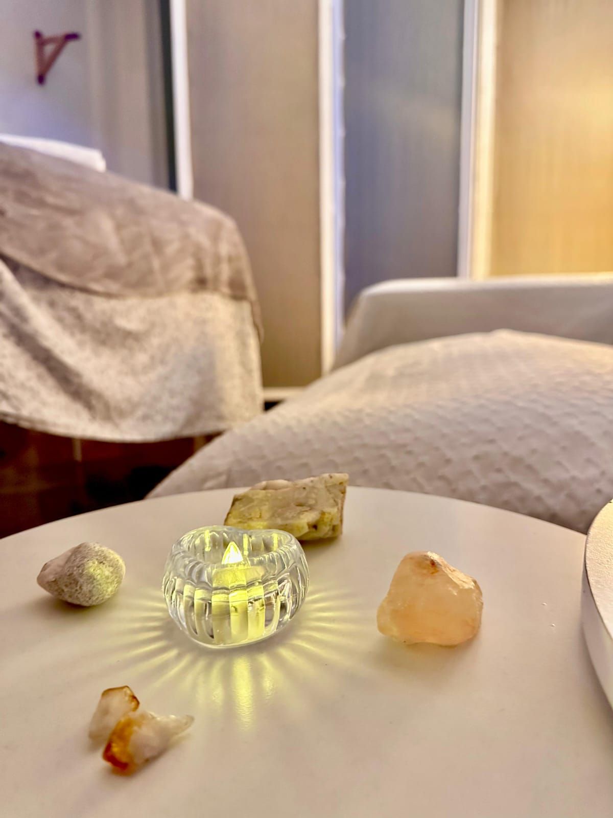 Pequeños cristales y un portavelas de vidrio sobre una mesa blanca en un dormitorio con luz tenue.