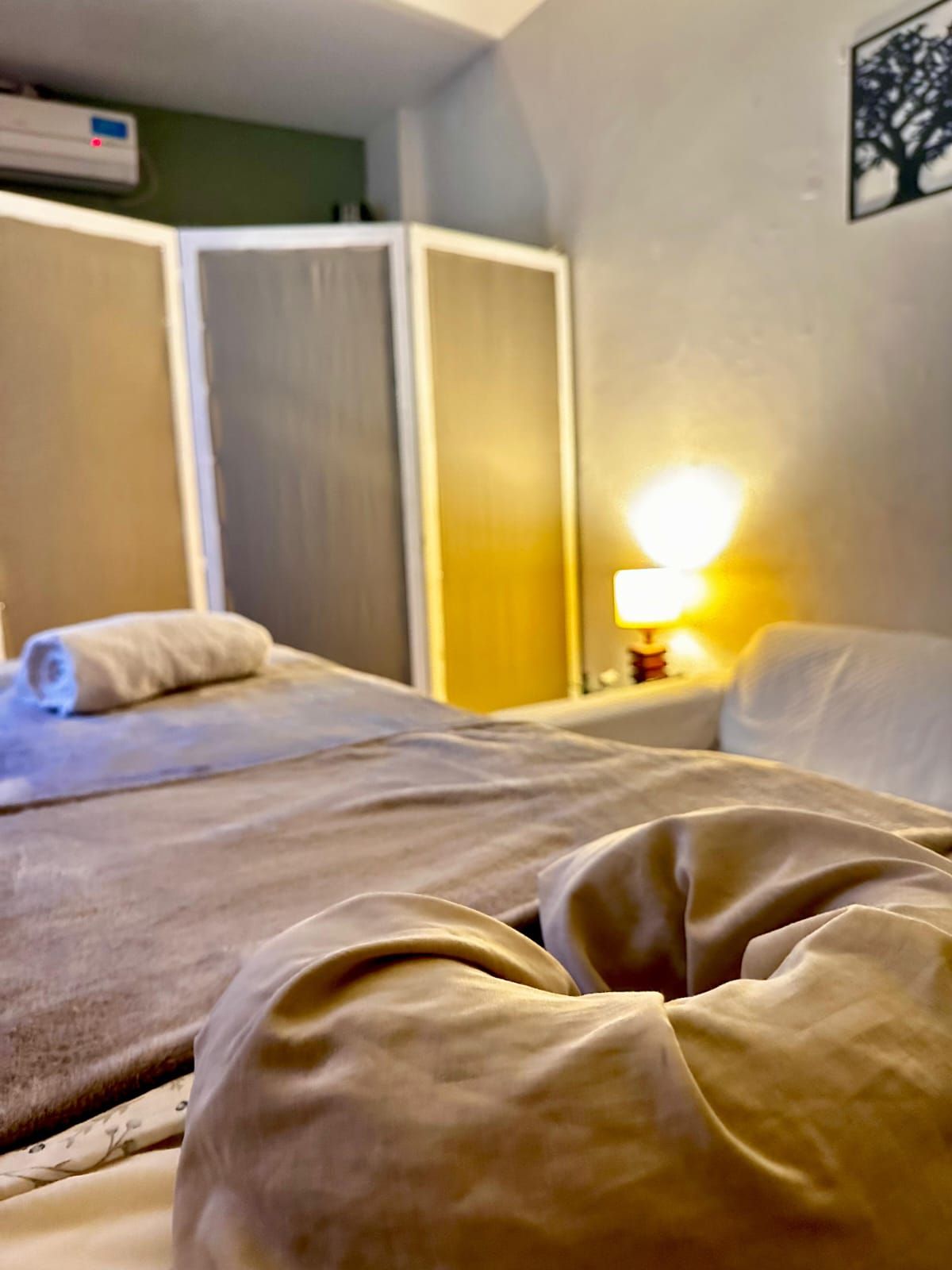 Dormitorio acogedor con cama, manta color tostado, biombo y la cálida luz de una lámpara de mesilla de noche