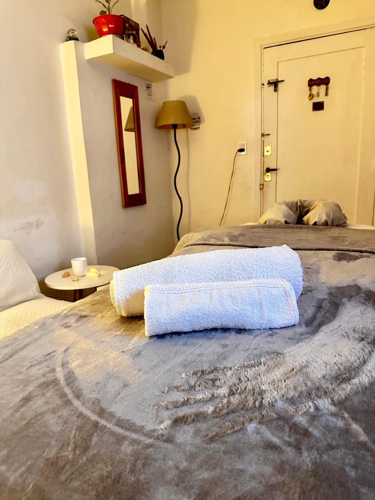 Acogedor dormitorio con colcha gris, toallas blancas dobladas, lámpara de mesita y espejo de pared