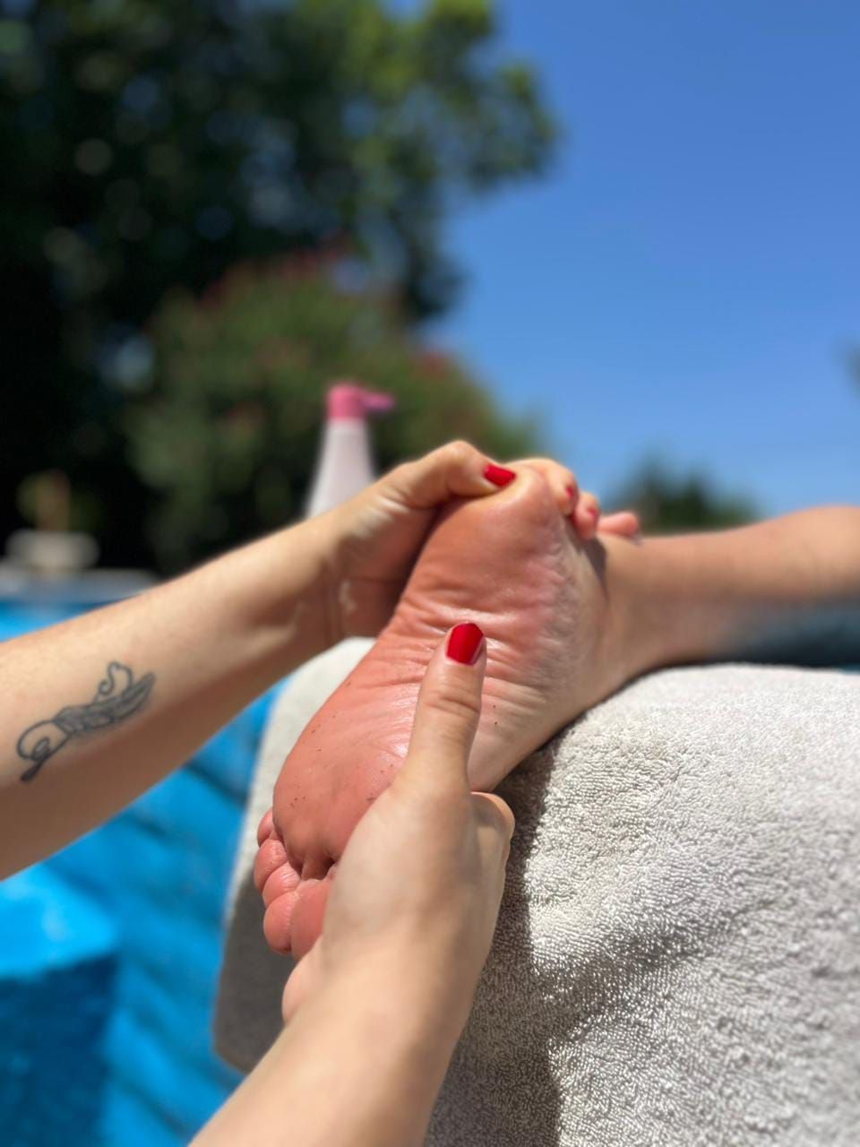 Manos sosteniendo un pie con las uñas pintadas de rojo junto a una piscina en un día soleado