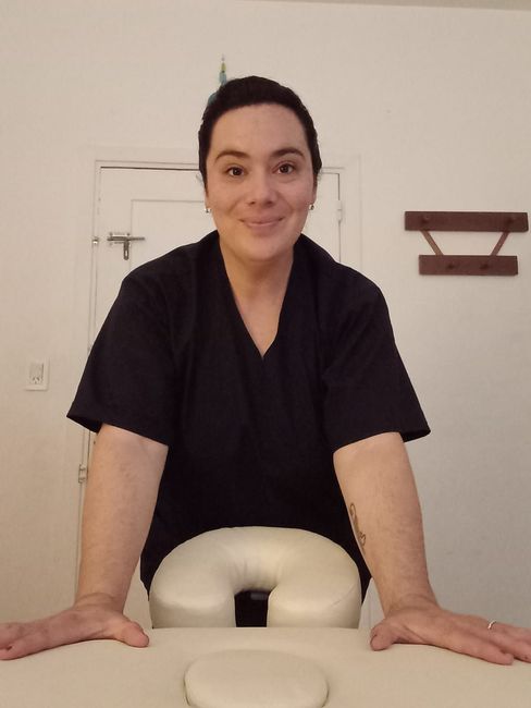 Persona con una camiseta negra apoyada en una camilla de masaje color crema, sonriendo en una sala de clínica.