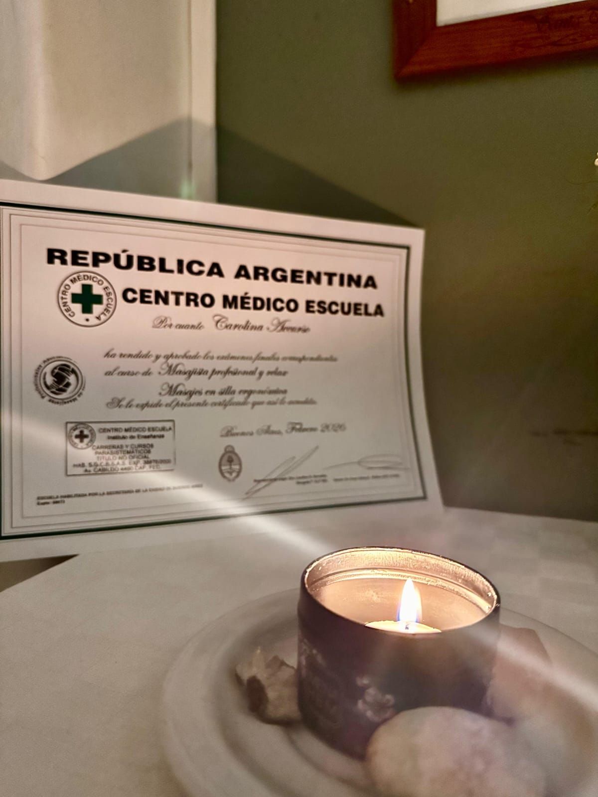 Vela encendida en un portavelas de vidrio, delante de un certificado de República Argentina Centro Médico Escuela enmarcado.