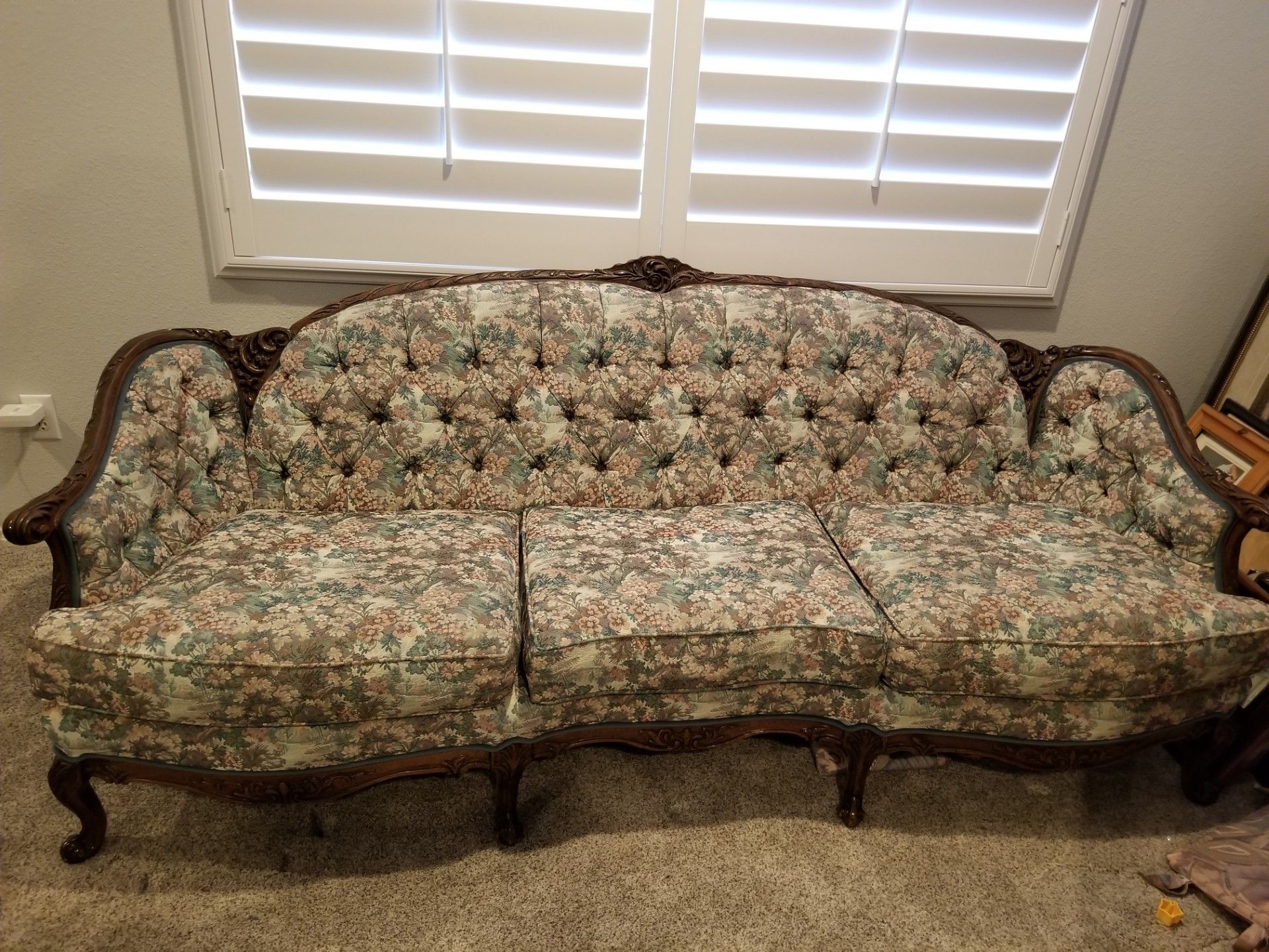 Floral-upholstered antique sofa with button tufting and dark wood trim, in front of shuttered window.