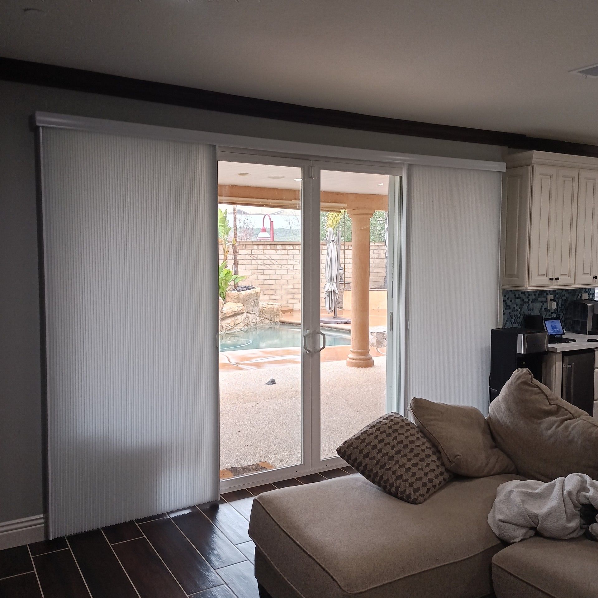 Sliding glass door with white panels, overlooking a pool and patio. Tan sofa, wood floor, and kitchen cabinets.