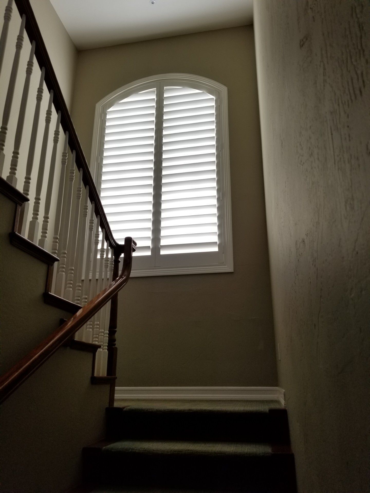 Staircase with wooden railing and white shutters on a arched window, in a tan-walled stairwell.