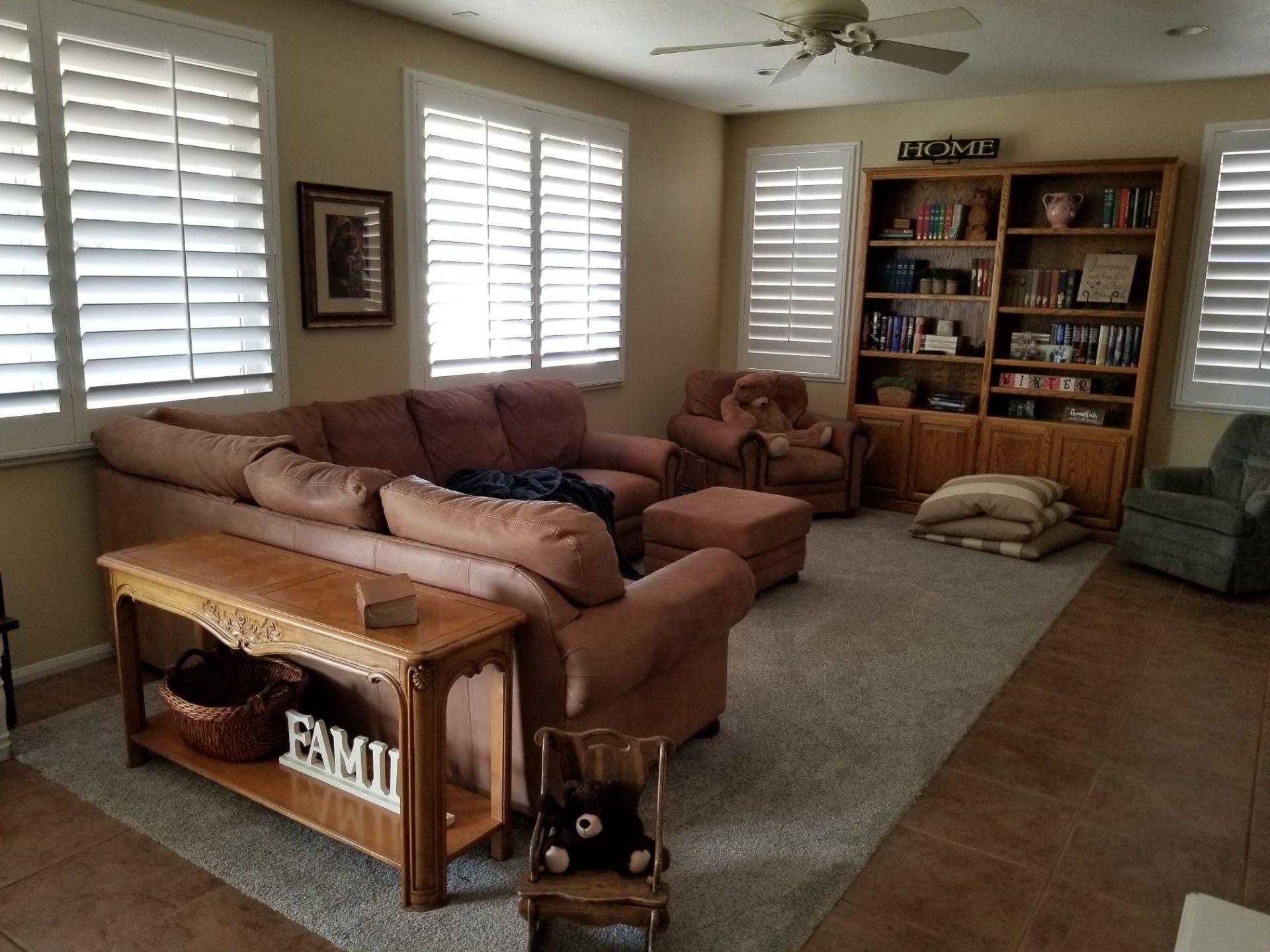 Living room with brown sectional sofa, wooden bookshelf, and windows with white shutters.