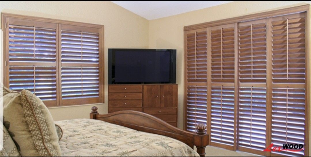 Bedroom with brown shutters on two windows and a TV over a dresser. Bed in the foreground.