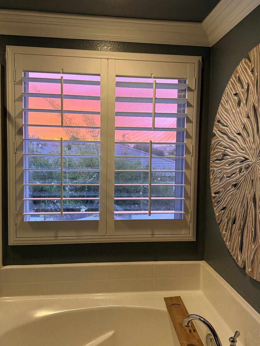 Bathroom window with shutters open to a colorful sunset, overlooking trees and a tub.