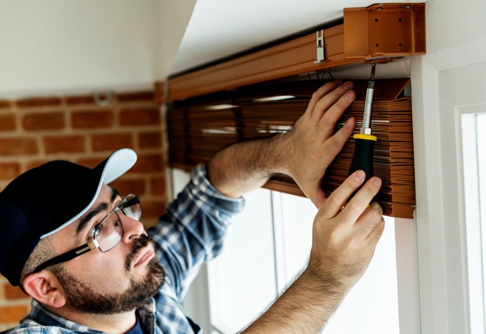 Person installing blinds, using a screwdriver, near a window with brick and white walls.
