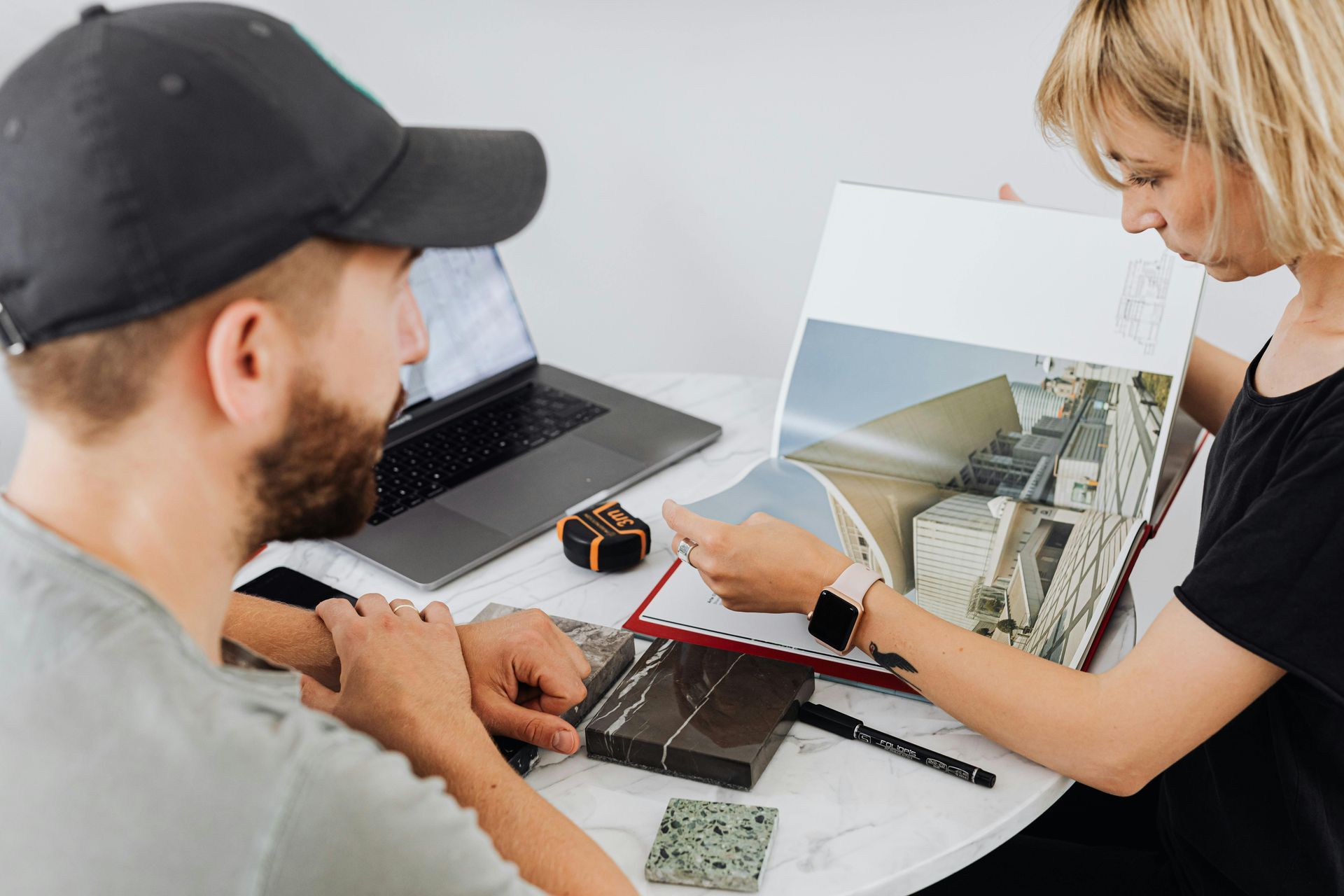 Person pointing to architectural design in a book, seated with a laptop and samples on a white table.