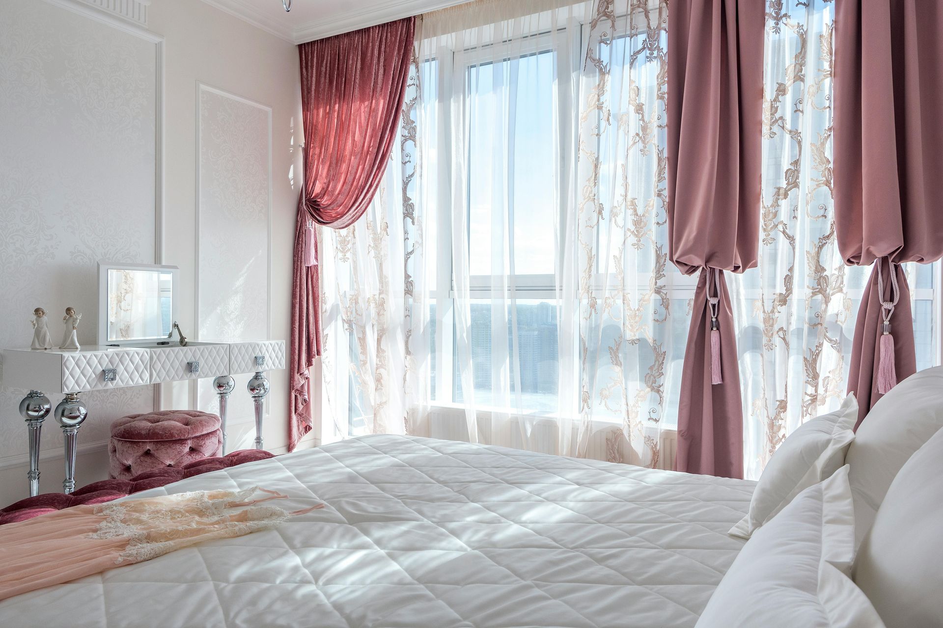 Bedroom with a large window, white bedding, and pink curtains framing a view. Vanity and stool on the left.