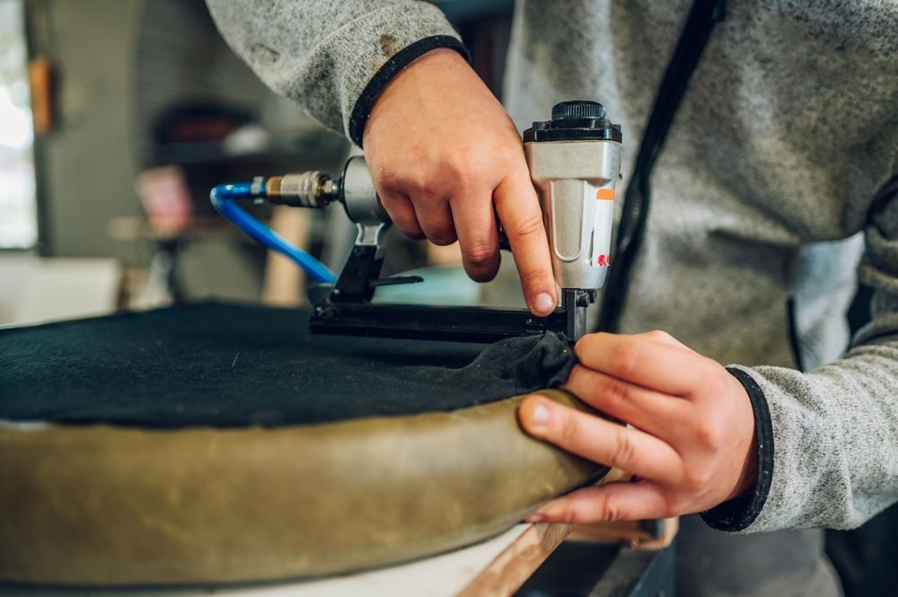 Person using a pneumatic staple gun to upholster a piece of furniture in a workshop.