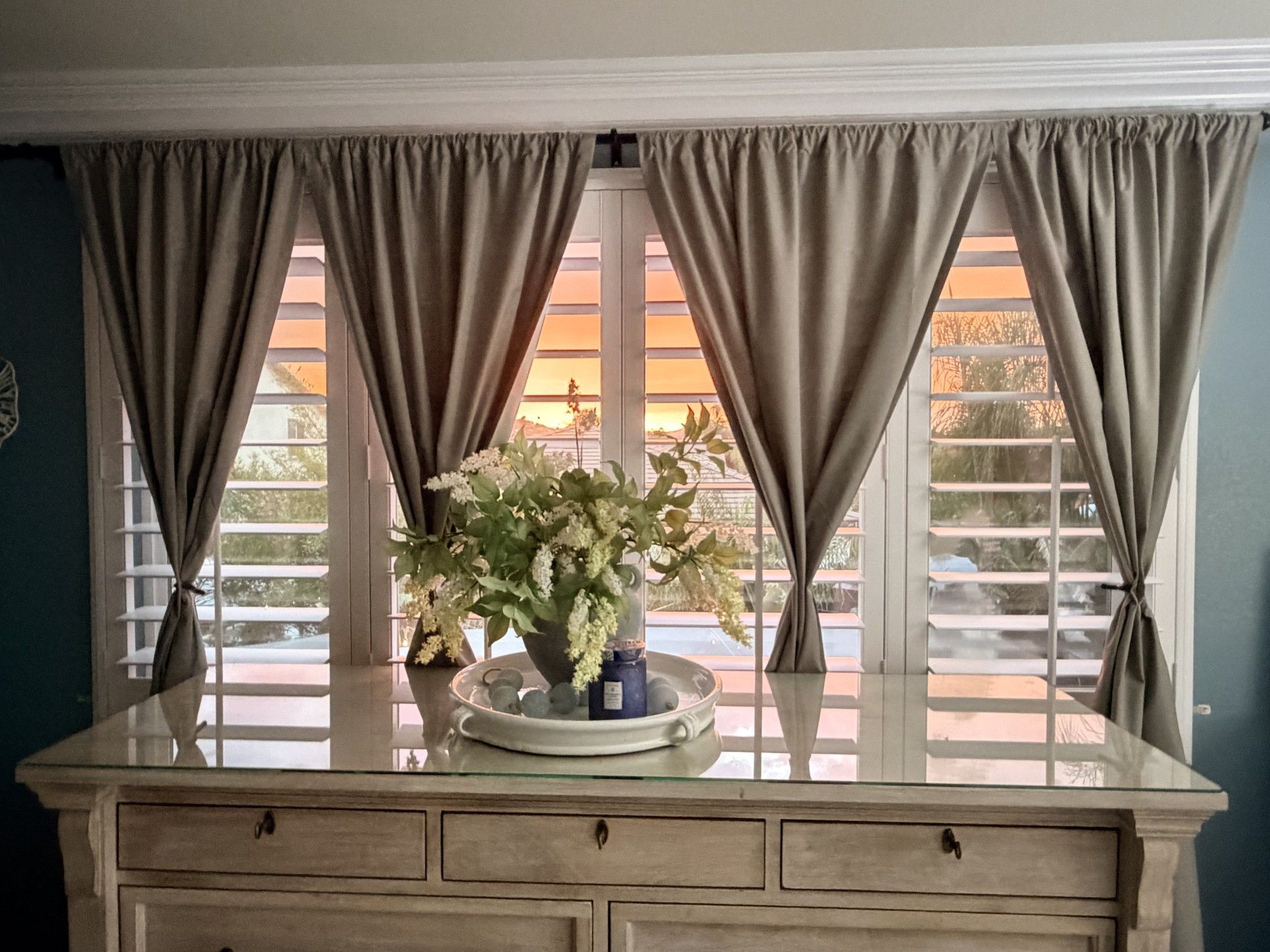 Drapes frame a window with shutters and a floral arrangement on a mirrored-top dresser. Warm sunset light.