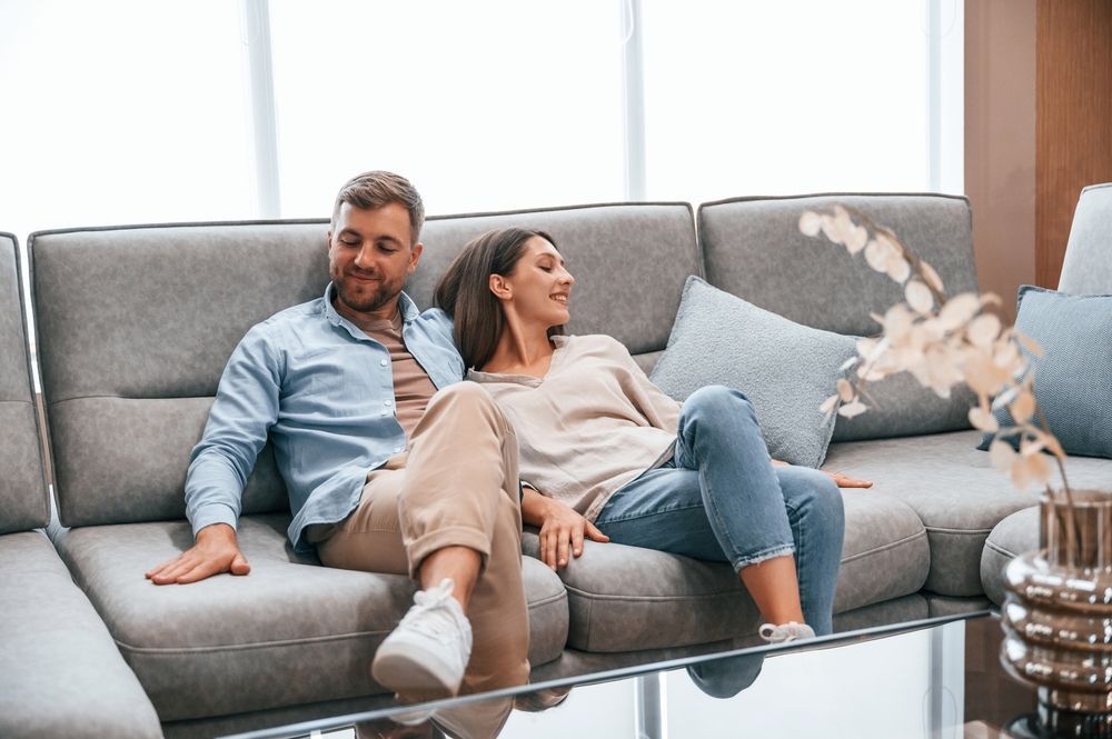 Couple relaxing on a gray sectional sofa, looking happy. Bright interior with a table and plant.