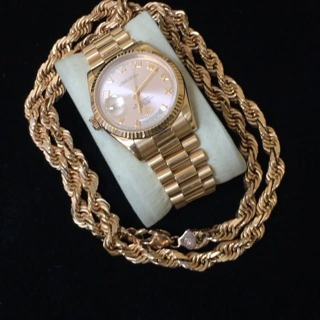 Gold wristwatch and braided gold chain displayed on a light blue cushion against a black background.