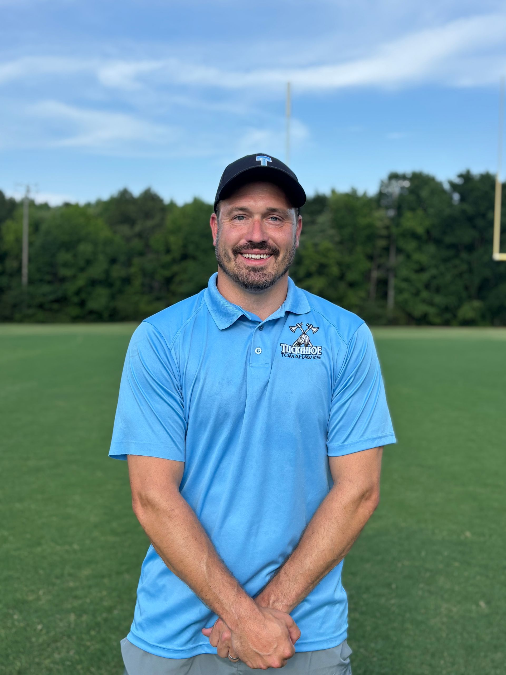 Football coach, arms crossed, on the field. Blue shirt, serious expression, players in background.