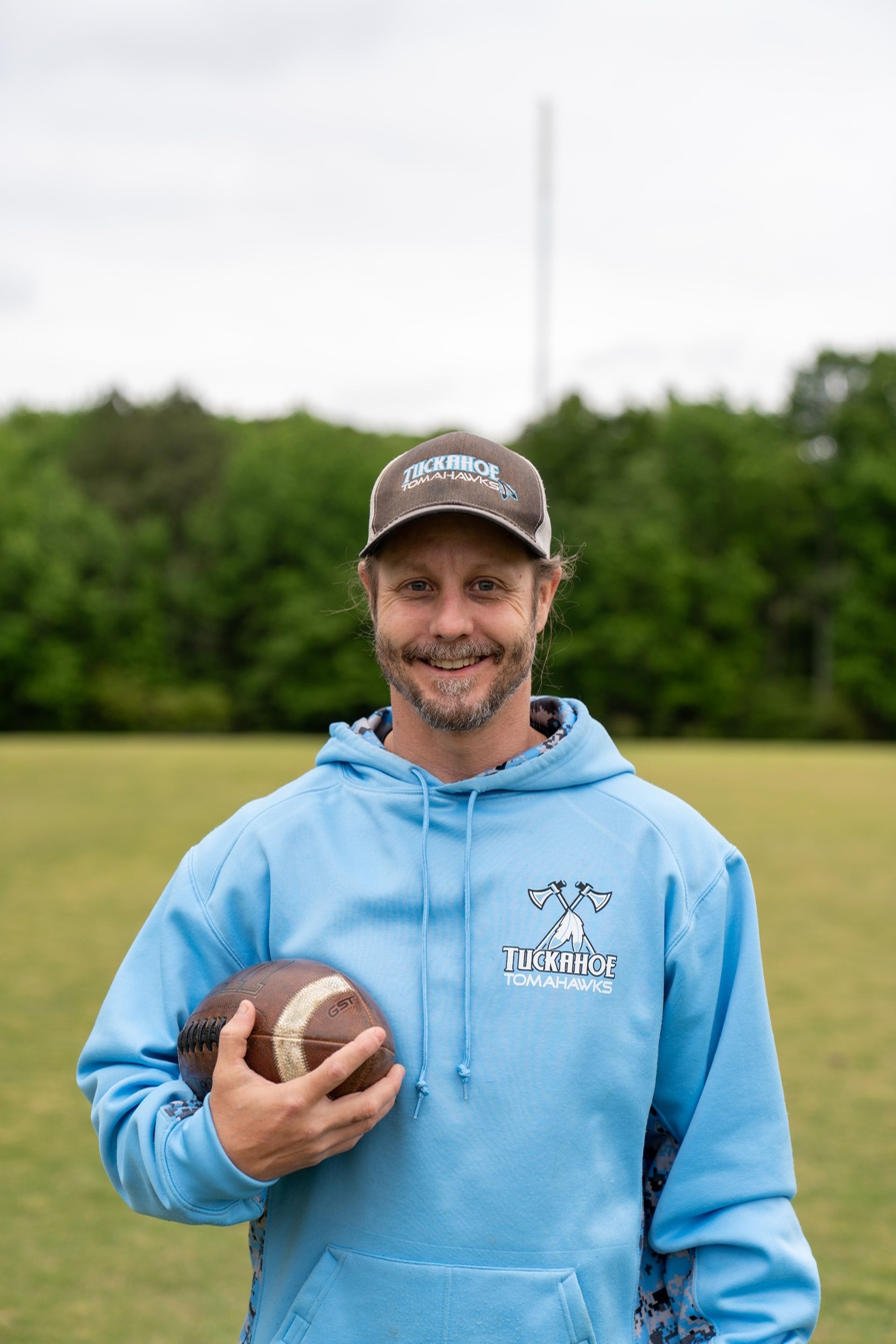 Man with crossed arms on a football field. He is wearing a blue polo shirt, khaki pants, and has a serious expression.