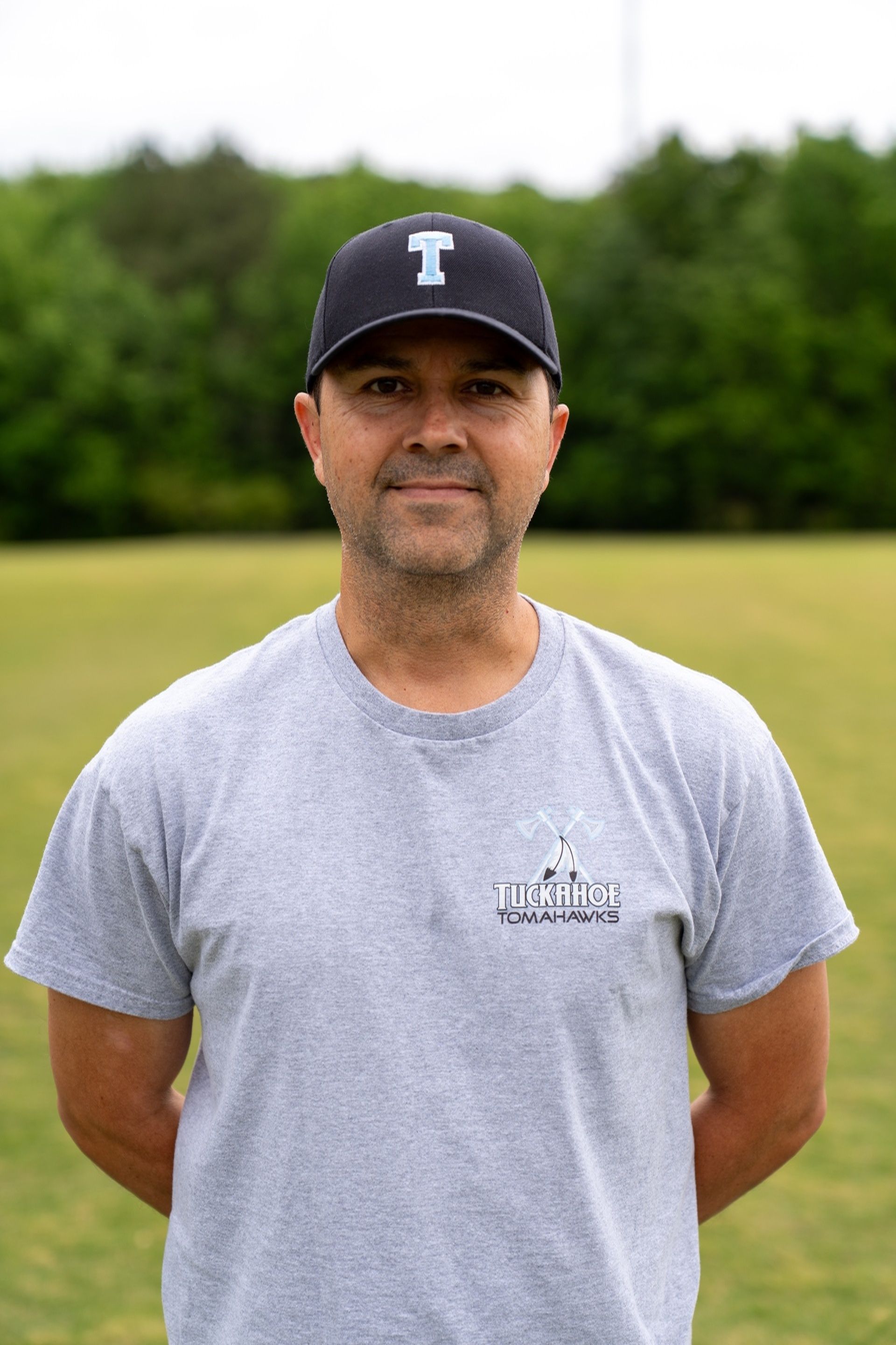 Man with crossed arms on a football field. He is wearing a blue polo shirt, khaki pants, and has a serious expression.