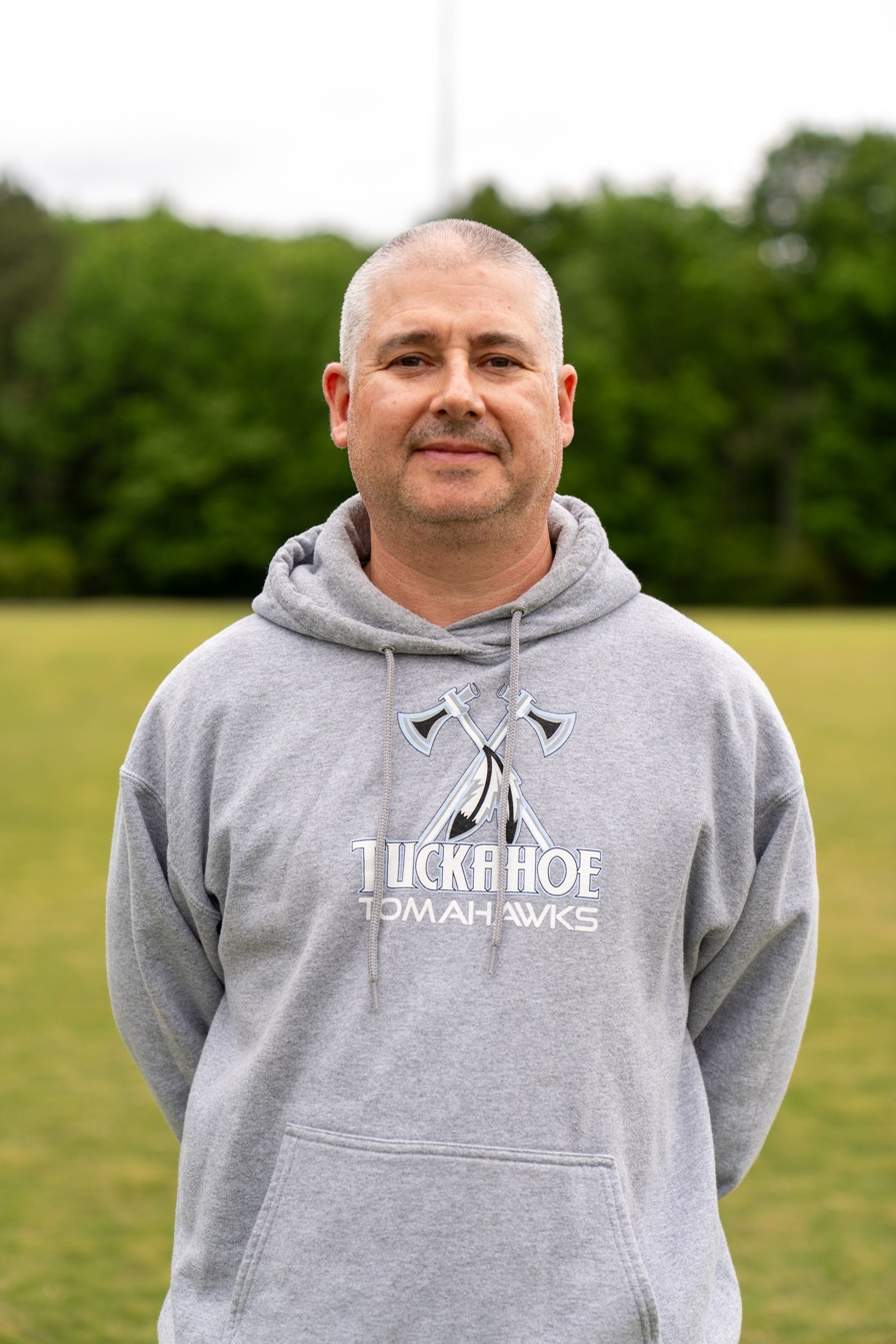 Football coach, arms crossed, on the field. Blue shirt, serious expression, players in background.