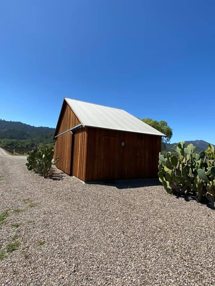 A brown wooden barn with a metal roof stands on a gravel path, surrounded by cacti under a clear blue sky.