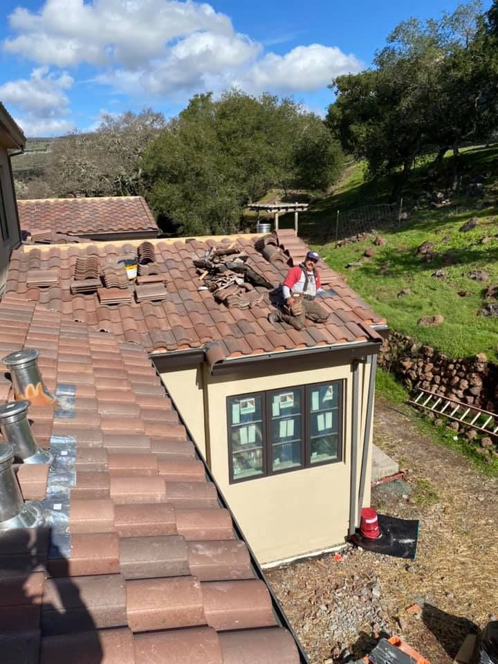 A person in a red shirt and work pants kneeling on a red-tiled roof, replacing tiles with a wooded background.