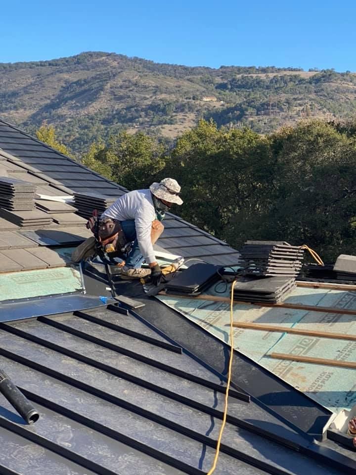 A worker in a sun hat repairs a dark-tiled roof on a sunny day, with a mountain and trees in the background.