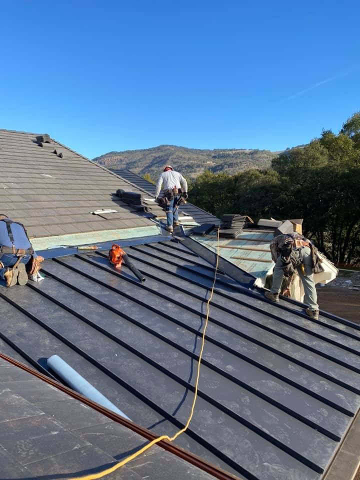 Workers installing a dark-colored metal roof on a house under a clear blue sky, with a mountain in the background.