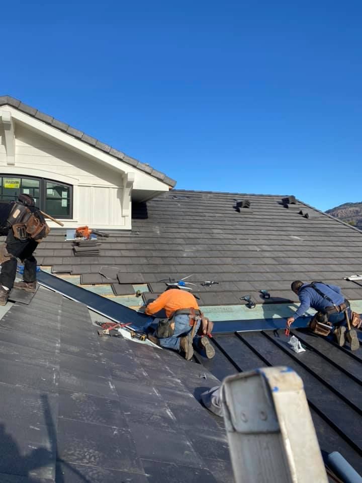 Three workers in safety gear repair a dark shingled roof on a sunny day with a clear blue sky.