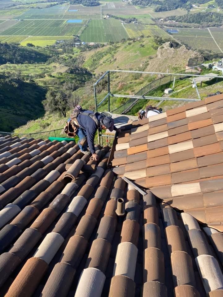 A worker in dark clothing repairs a sloped, terracotta-tiled roof overlooking a valley with fields and hills.