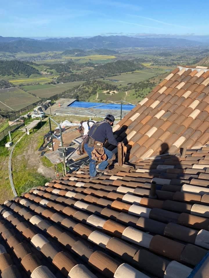 A person wears a tool belt while installing brown, barrel-style tiles on a steep roof overlooking a scenic valley.