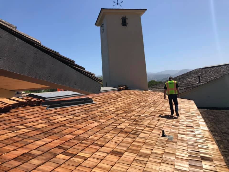 A worker in a high-visibility vest walks across a newly installed cedar shake roof toward a tall, light-colored tower.