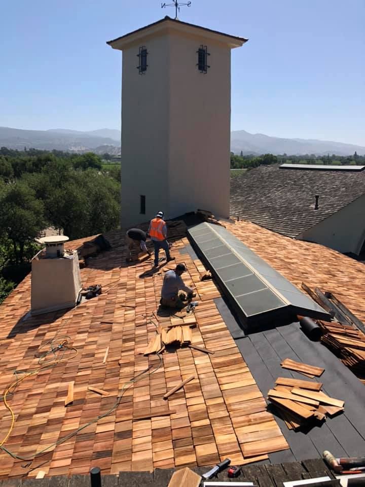 Three workers in high-visibility vests install cedar shake shingles on a large pitched roof near a bell tower.