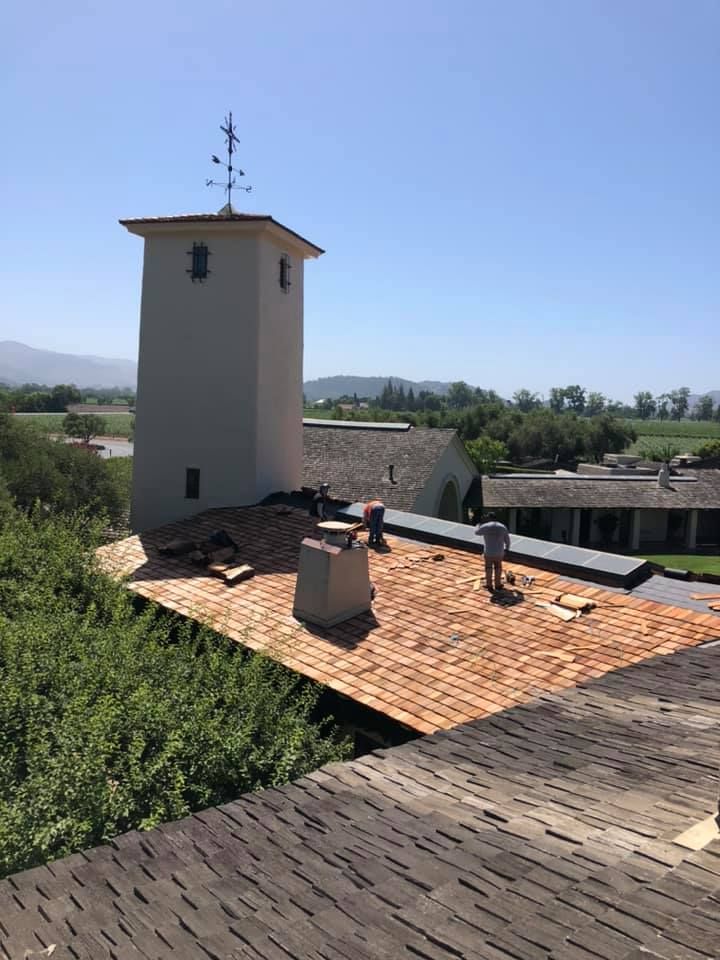 Construction workers on a cedar shake roof near a tower, with a view of hills and trees in the background.