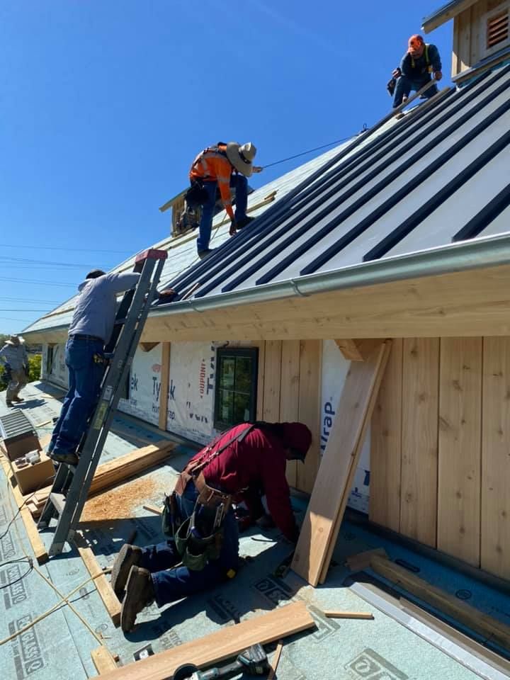 Four workers in work clothes install metal roofing and exterior siding on a new building under a clear blue sky.