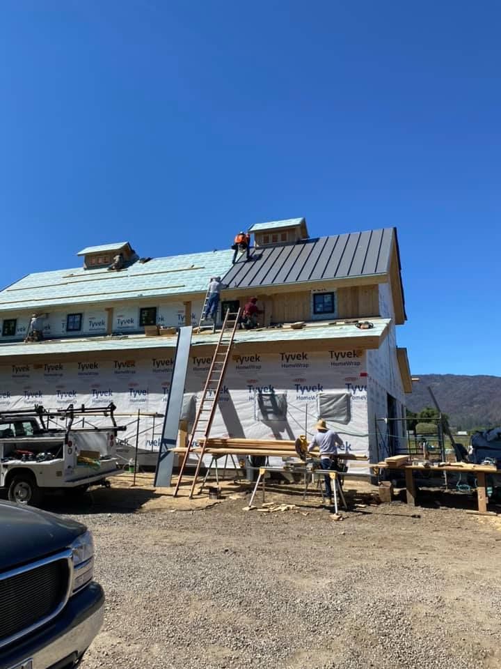 Workers install a metal roof on a building under construction on a sunny day with mountains in the background.