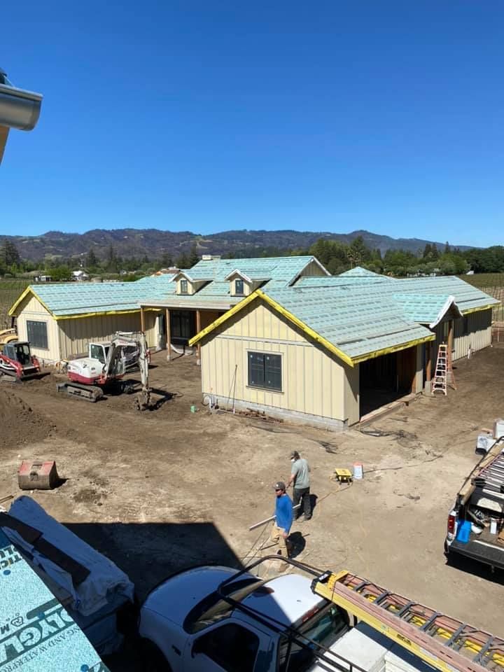 Workers at a construction site with a large, partially completed house featuring green roof sheathing and yellow siding.