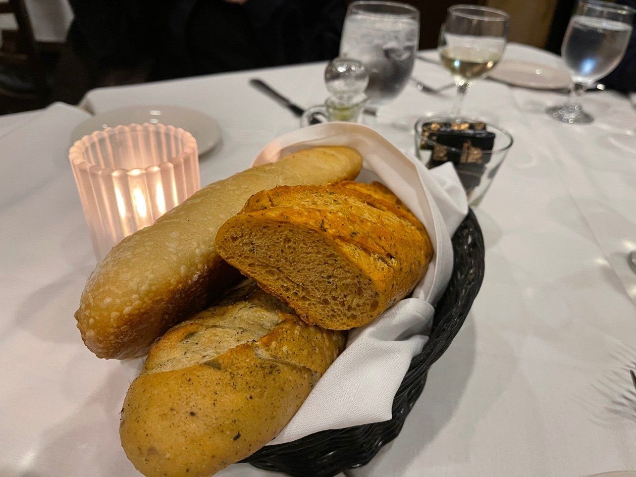 Basket of bread on a table, including baguette and rolls, next to a lit candle and glasses of water and wine.