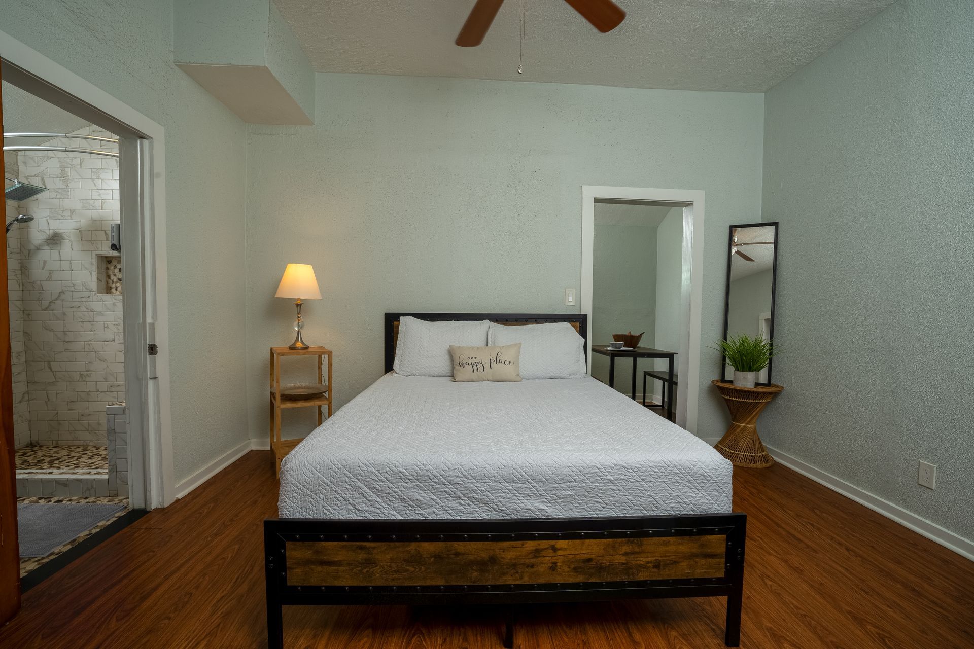 Bedroom with bed, nightstand, mirror, and a bathroom entrance. Light blue walls, wood floor.