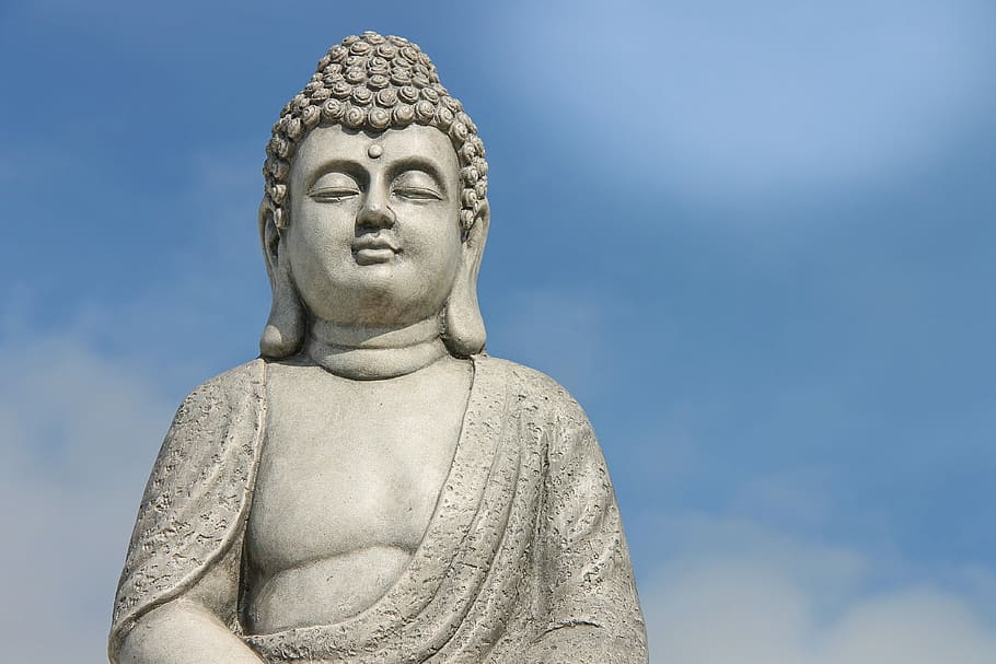 Statue of Buddha with closed eyes, draped in robes, against a blue sky with clouds.