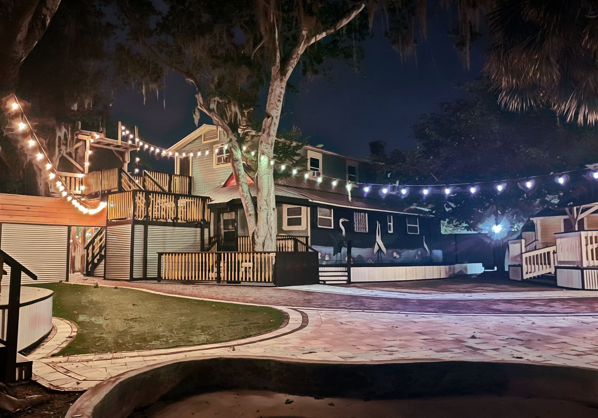 Night view of a multi-level outdoor venue with string lights, a grassy area, and a building.