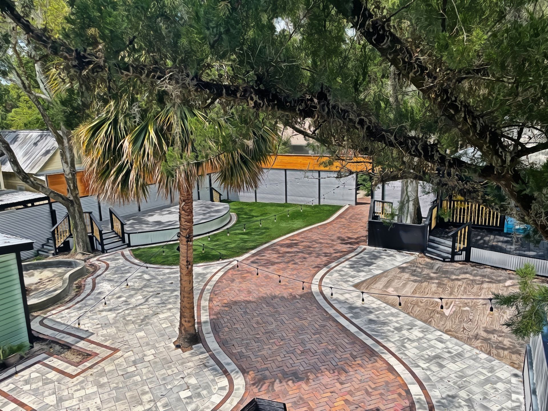 Brick path winds through a courtyard with greenery, a palm tree, and buildings in the background.