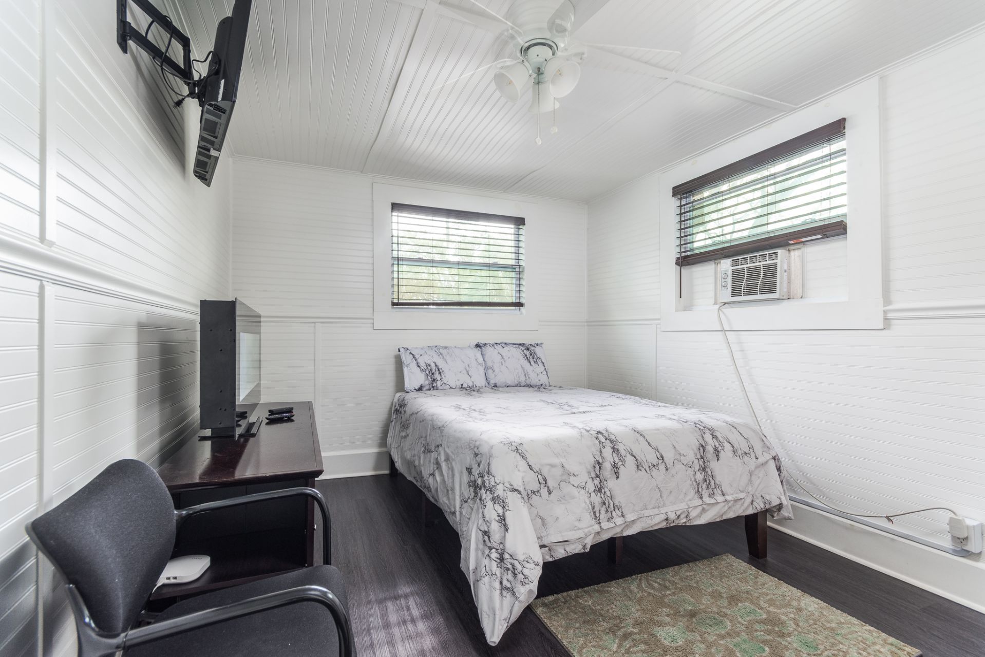 Small bedroom with a bed, TV, chair, and a window with blinds; white walls, patterned ceiling.