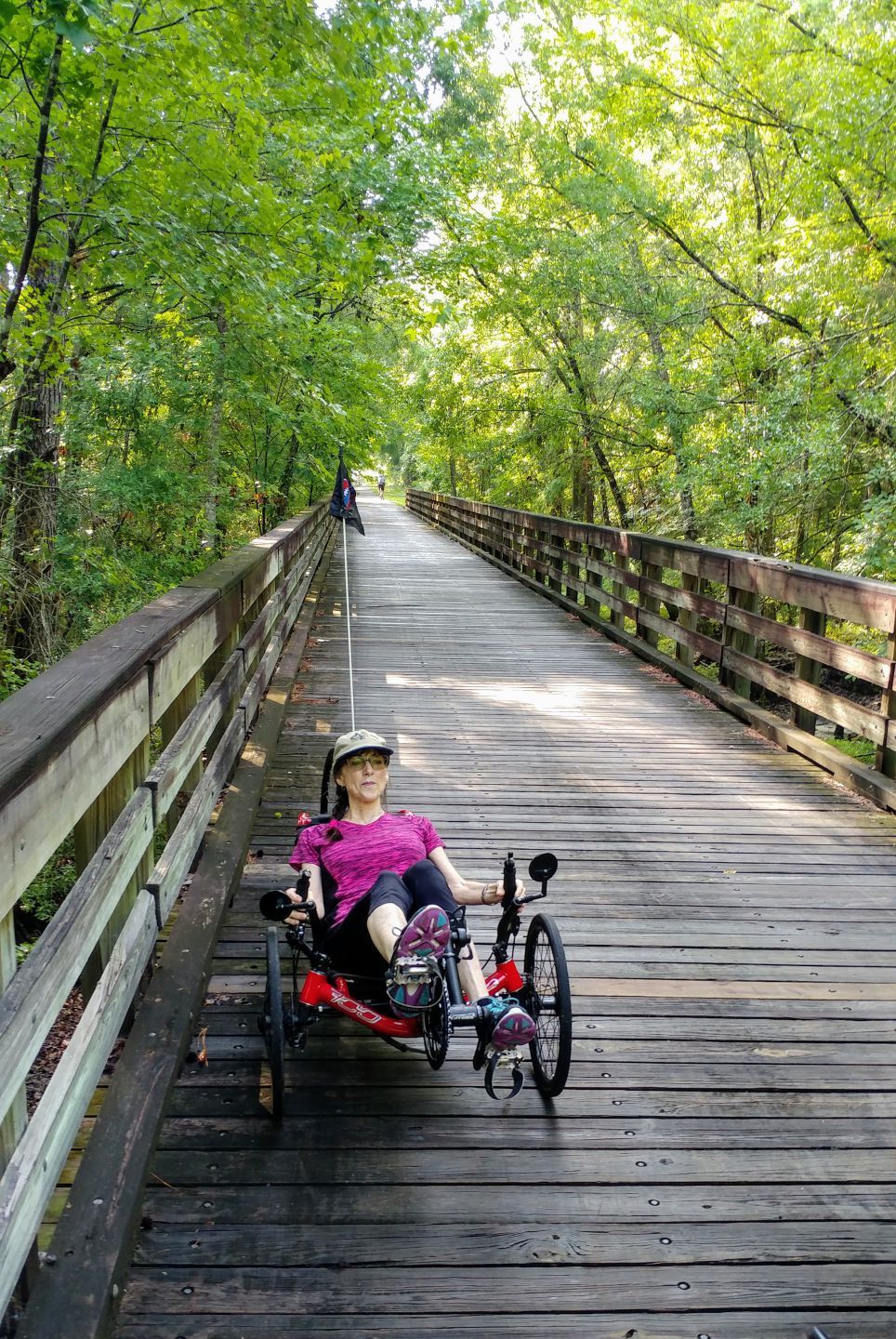 Woman on recumbent trike rides across wooden bridge lined with trees.