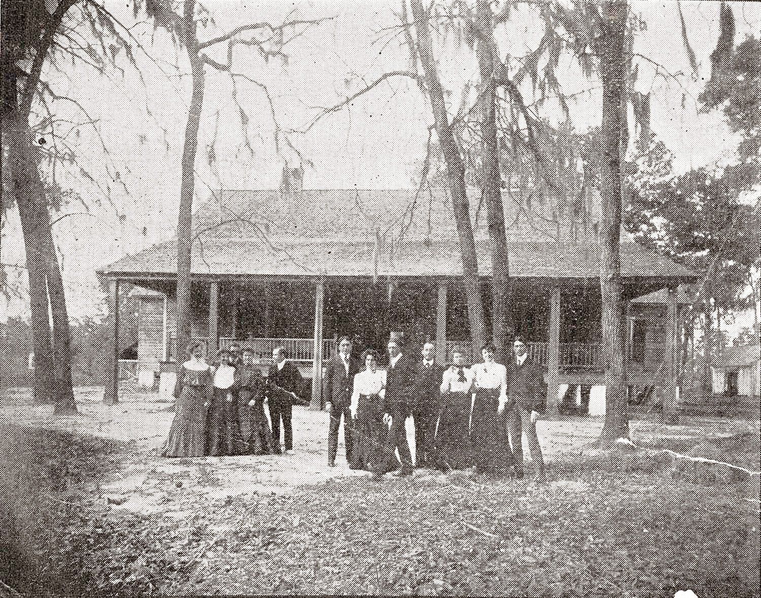 Group of people standing in front of a building in a wooded area. Black and white photograph.