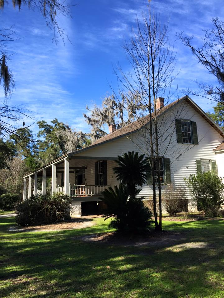 White house with porch, green shutters, nestled among trees, under a blue sky.